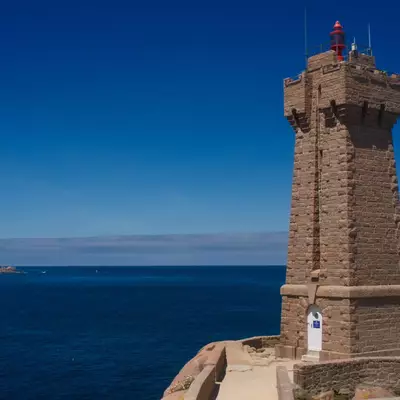 A stone tower lighthouse stands on a rocky coastal outcrop overlooking the Mediterranean Sea under clear blue skies. The cylindrical tower features crenellations at its top and a small shrine or marker at its base. The coastline and distant shoreline are visible across the calm blue water.