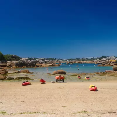 A sandy beach with large boulders scattered throughout, shallow turquoise water, and red kayaks lined up on the shore under a clear blue sky, with residential buildings visible along the coastline in the background.