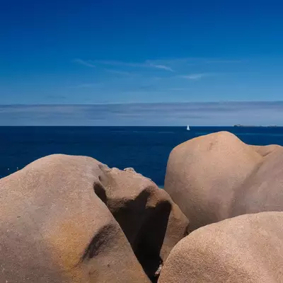 Large smooth granite boulders in shades of tan and cream sit against a deep blue ocean and clear sky, with a sailboat visible on the horizon.