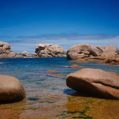A pristine coastal cove with crystal-clear turquoise water surrounded by massive rounded granite boulders under a bright blue sky. The rocky shoreline features smooth, weathered stone formations in various sizes, with patches of green algae visible in the shallow water. This scenic beach exemplifies the dramatic granite landscape characteristic of the Breton coast.