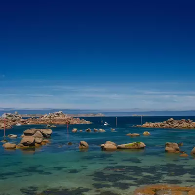A pristine Atlantic cove with turquoise water scattered with large granite boulders, rocky outcrops, and a sailboat, under a clear blue sky on the Sentier des douaniers coastal path.