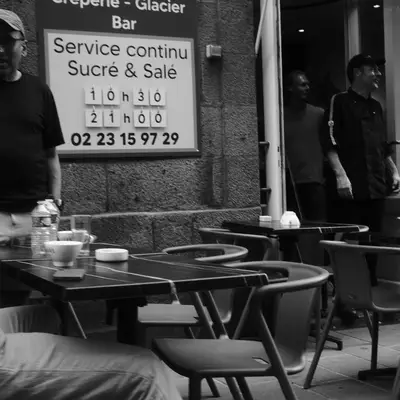 A black and white street scene of an outdoor café in St-Malo with empty tables and chairs in the foreground. A man in a dark shirt and cap stands near the storefront, while other figures are visible in the background. A sign on the window advertises 