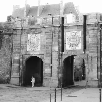 Historic stone fortification gate with two large arched entrances and decorative heraldic shields mounted on the facade, located in St-Malo's walled city. The black and white photograph shows the imposing medieval architecture with crenellated towers above the gatehouse and a courtyard area in front.