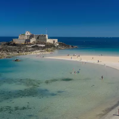 A medieval fortress sits on a rocky island surrounded by turquoise shallow waters and a pristine sandy beach filled with visitors in St-Malo, France. The clear blue sky and calm sea create ideal beach conditions, with the fortified structure dominating the landscape.