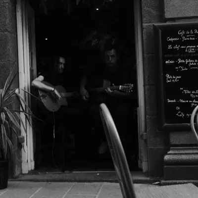 A black and white photograph of a storefront entrance in St-Malo with a dark doorway, potted plant on the left, menu board on the right, and a metal chair visible in the foreground. The scene captures the intimate atmosphere of what appears to be a small French restaurant or café.