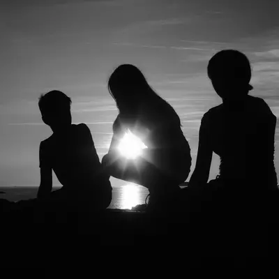 Three silhouetted figures sit together against a dramatic sunset or sunrise over water, with bright light creating a glowing effect between them. The black and white composition emphasizes their outlines and the luminous sky with clouds above.
