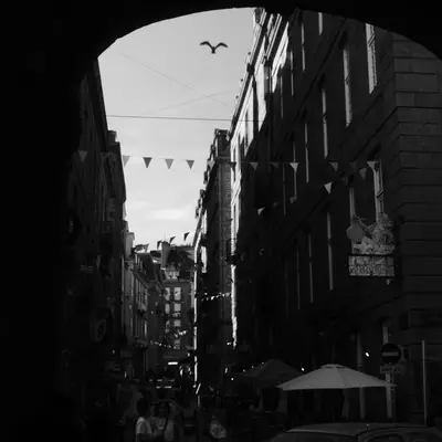 A narrow European street lined with tall buildings frames a distant view of sky and a flying bird, with decorative bunting strung overhead and a café umbrella visible below, photographed in black and white.