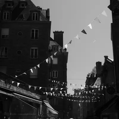 A narrow medieval street in St-Malo lined with tall stone buildings, strung with white triangular bunting flags overhead, photographed in black and white with a castle tower visible in the distance.