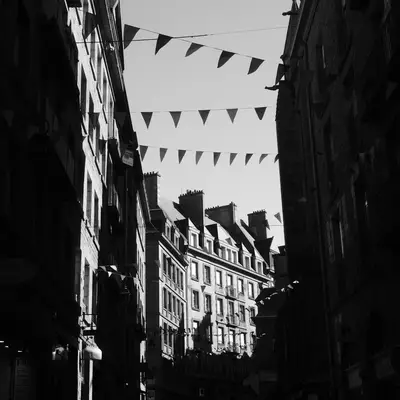 A narrow European street lined with historic buildings framed by dark facades, with decorative bunting strung overhead against a light sky. The black and white photograph captures the architectural character and festive atmosphere of what appears to be a medieval town street.