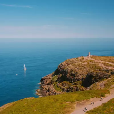 A scenic coastal landscape showing a dramatic headland with green grassland and rocky cliffs overlooking turquoise waters, with a white lighthouse visible on the peninsula and a sailboat in the distance below.