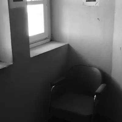 A sparse interior room with a black armchair positioned against a white wall, a small window with bright natural light on the left, and a white electrical outlet on the right wall.