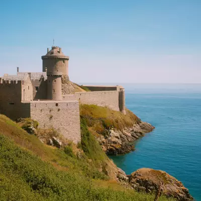 A stone fortress with a round tower stands on a rocky coastal cliff overlooking turquoise waters under a clear blue sky. Green grass covers the sloping terrain leading down to the sea, with the fortified structure featuring medieval architecture and defensive walls.