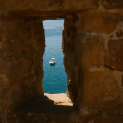 A boat on turquoise water viewed through a narrow rectangular stone opening in an ancient fortress wall at Cap Fréhel, Fort La Latte.
