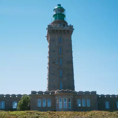 A tall stone lighthouse with a green lantern room at the top stands behind a low fortified building with blue-shuttered windows, set on a grassy hillside under clear blue sky.