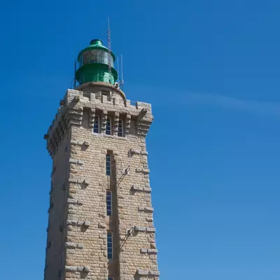 A tall stone lighthouse with a green lantern room at its top stands against a clear blue sky. The cylindrical tower features narrow windows and appears to be a historic maritime structure. The architectural style and construction suggest it's located on the French coast near Cap Fréhel.