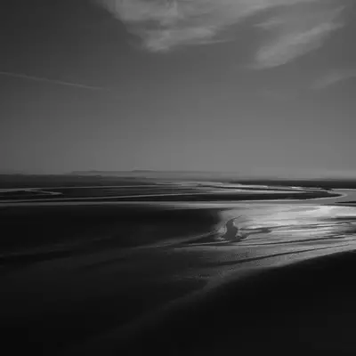 A black and white coastal landscape showing a vast tidal flat with winding water channels reflecting light, under an overcast sky with subtle clouds on the horizon.