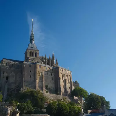 Mont Saint-Michel's iconic abbey crowns a rocky tidal island in Normandy, France, with its Gothic spire rising prominently against a clear blue sky. Stone fortification walls and buildings cascade down the granite mount, surrounded by lush green vegetation at its base.