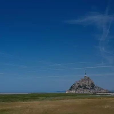 Mont-Saint-Michel rises dramatically from a vast expanse of tidal flats and green meadows under a clear blue sky. The medieval abbey and fortified village crown the rocky tidal island, its distinctive silhouette visible across the flat Normandy landscape.