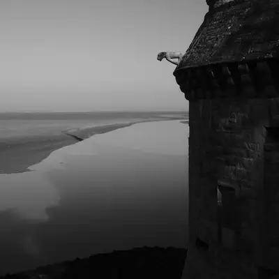 A stone watchtower with a domed top stands in the foreground of this black and white photograph, overlooking a vast tidal bay with winding waterways below. The dramatic coastal landscape stretches to a distant horizon under an overcast sky.