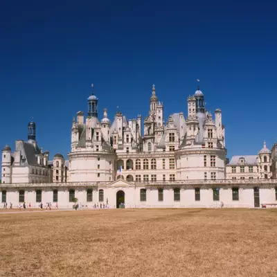 A grand French Renaissance château with distinctive white limestone towers and ornate architectural details stands against a clear blue sky, viewed from a large sandy courtyard in front of the main façade.
