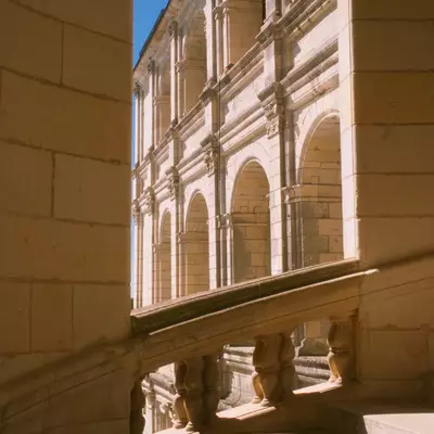 A Renaissance palazzo facade framed through a stone archway, featuring multiple levels of arched windows and classical columns bathed in sunlight, with shadows cast on an interior floor in the foreground.