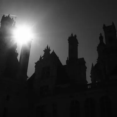 Silhouetted Gothic castle towers and spires against a bright sun breaking through overcast sky, creating a dramatic high-contrast black and white scene of an imposing medieval fortress.