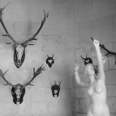 A white-painted figure with raised arms stands against a white brick wall surrounded by mounted animal skulls and horns, including deer antlers and various horned creatures arranged in a symmetrical pattern.