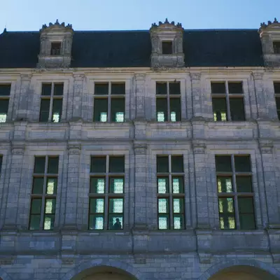 A symmetrical stone building facade with three mansard roofs topped with ornamental chimneys, featuring two rows of tall rectangular windows with green frames arranged in a regular pattern across the light-colored stone exterior.