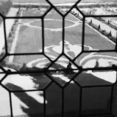 Black and white photograph taken through a chain-link fence showing agricultural fields with curved rows and a tree line in the distance under an overcast sky.