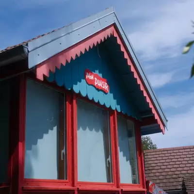 A red and blue wooden kiosk with a decorative scalloped awning sits against a partly cloudy sky, with evergreen branches visible on the right side and another building's roof visible in the background.