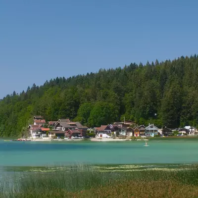 A turquoise alpine lake fronts a cluster of chalets and buildings nestled against a densely forested hillside of evergreen trees under clear blue sky.
