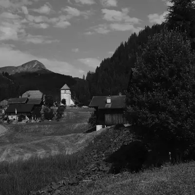A black and white landscape photograph of a rural mountain valley with a small wooden church, scattered buildings, and evergreen forests under a partly cloudy sky, with a snow-capped peak visible in the distance.