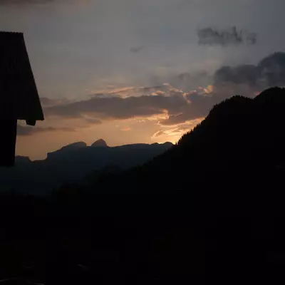 A dramatic sunset breaks through clouds over silhouetted mountains and forested ridges, with a dark building structure visible on the left side of the frame.