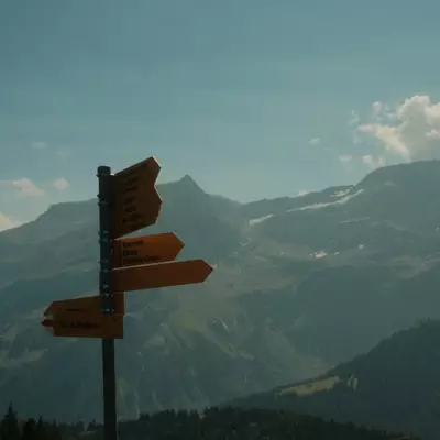 A wooden directional signpost with multiple arrow signs points toward mountain trails, with layered Alpine peaks and forested slopes visible in the background under a partly cloudy sky.