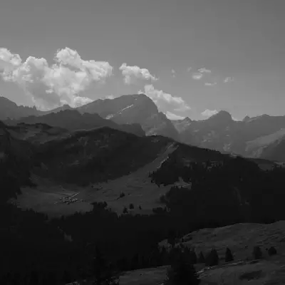 Dramatic black and white mountain landscape with rolling hills in the foreground, a valley with scattered buildings nestled below, and jagged Alpine peaks rising against a cloudy sky in the background.