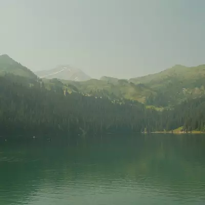 A serene mountain lake with emerald-green water surrounded by dense coniferous forests and distant snow-capped peaks under an overcast sky. A small red boat is visible on the calm water near the left shore.