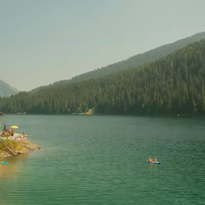 A turquoise Alpine lake surrounded by dense coniferous forest and mountains, with a small wooden cabin on the left shore and a few people swimming in the calm water.