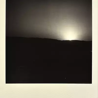 Sun setting or eclipsing behind dark silhouetted hills with luminous clouds in a grainy polaroid-style photograph.