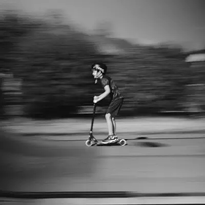 A young skateboarder rides a scooter across pavement with motion blur surrounding them in black and white.
