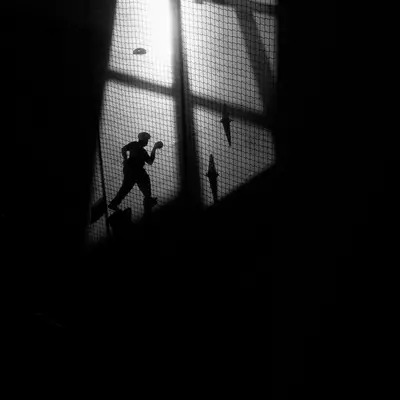 Silhouette of a person running through a bright doorway, framed by dark netting and diagonal shadows.