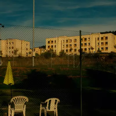 Two empty white plastic chairs and a folded yellow umbrella behind a chain-link fence, with apartment buildings beyond.
