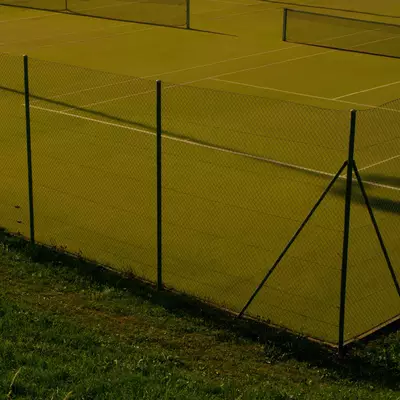 Chain-link fence surrounding a tennis court, bathed in yellow-green light with long shadows cast across the surface.
