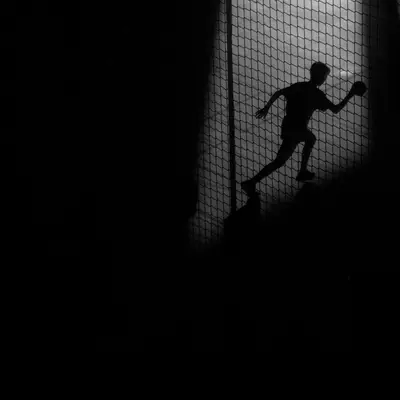 Silhouette of a young handball player caught mid-throw, backlit against a mesh fence in near darkness.