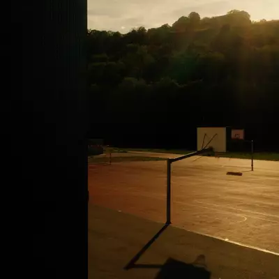 Silhouette of a basketball hoop on an outdoor court at sunset, with a dark building edge framing the left side of the scene.