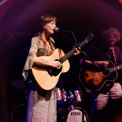 Female singer playing an acoustic guitar in front of a microphone, accompanied by a guitarist, on stage with a circular vignette effect.