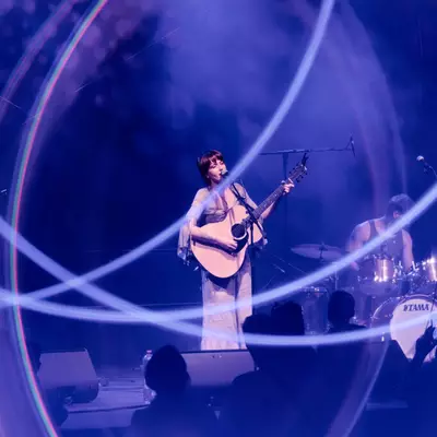 Female musician playing guitar on stage, lit by circular purple and blue light halos, with a drummer in the background.