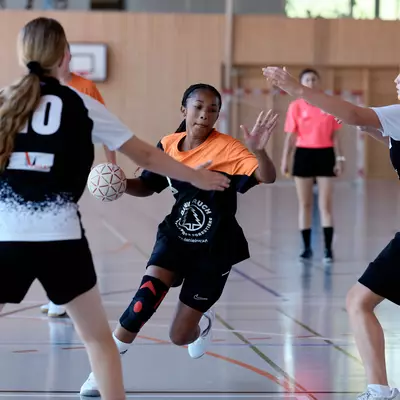 A female player in orange carrying the ball, flanked by two defenders in black and white on a handball court.