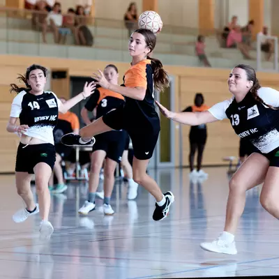Female player in an orange jersey jumping to take a shot at goal, surrounded by two opponents in white during a women's handball match.