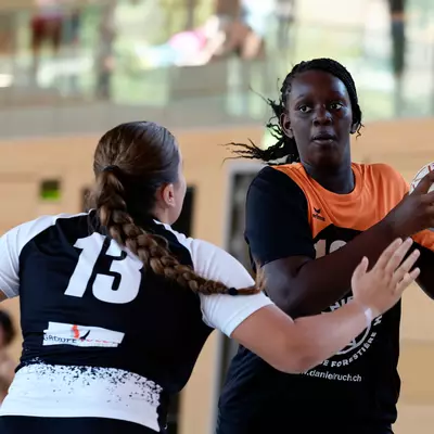 Handball player in an orange jersey shielding the ball from a defender wearing the number 13.