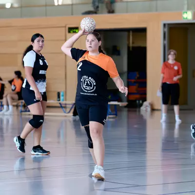 Handball player in an orange and black kit preparing to shoot, ball raised, on an indoor court.